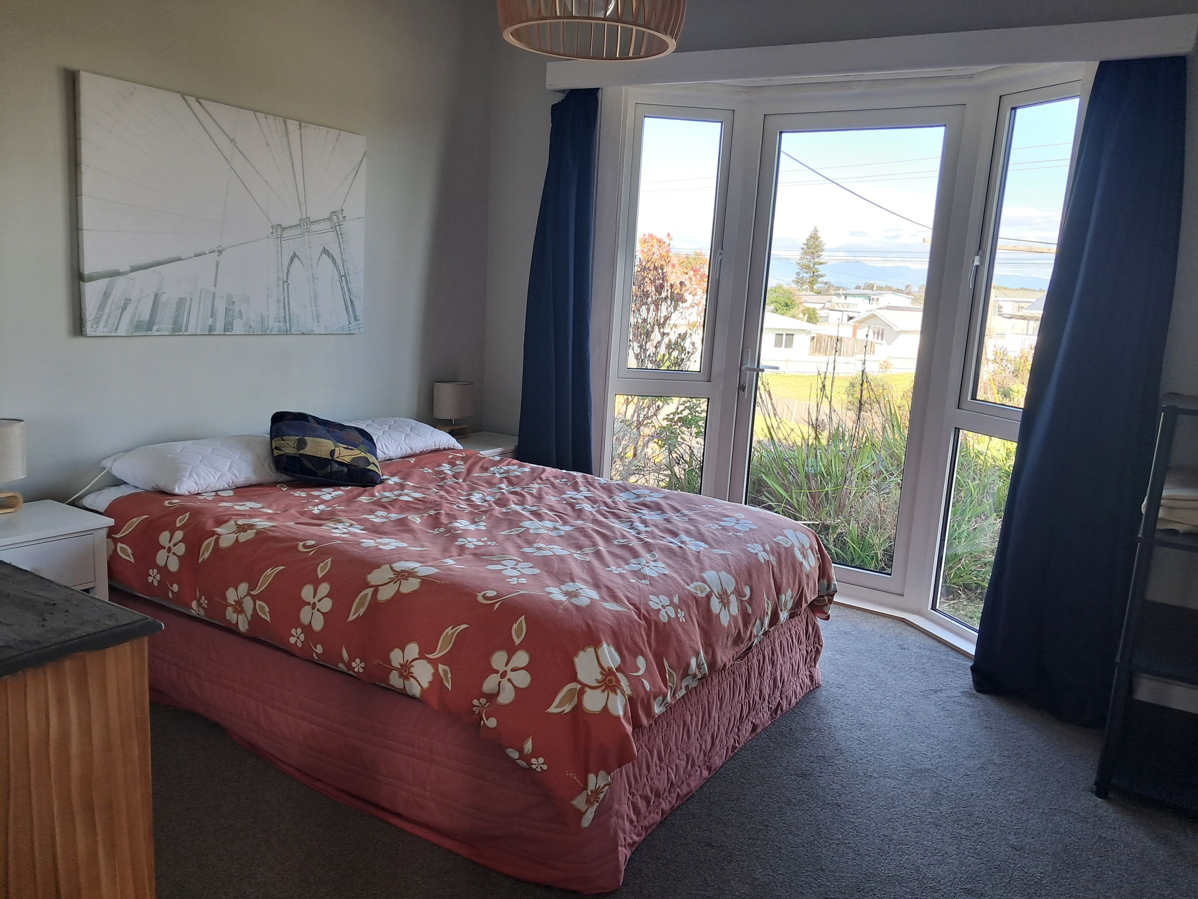 Un dormitorio con una cama, una mesita de noche, una lámpara, una ventana con cortinas y una obra de arte colgada en la pared.