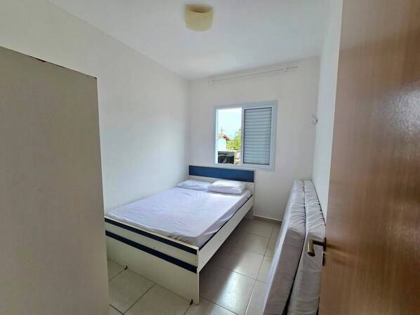 Un dormitorio con una cama individual, una ventana con persianas, una puerta y un pequeño banco.