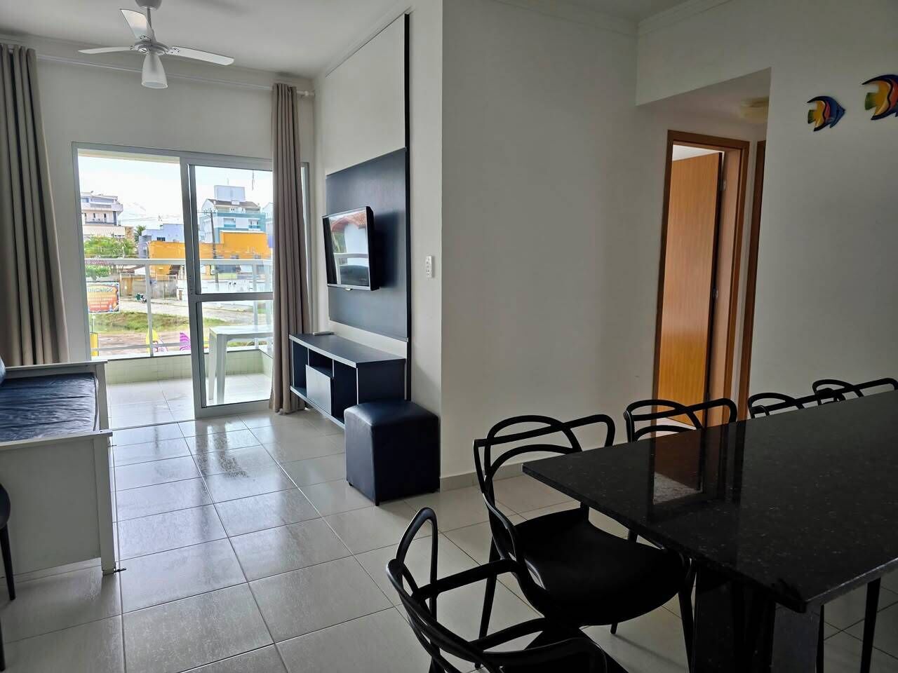 Een moderne woonkamer met een flatscreen televisie, een eettafel met stoelen, een plafondventilator en een schuifpui naar een balkon.