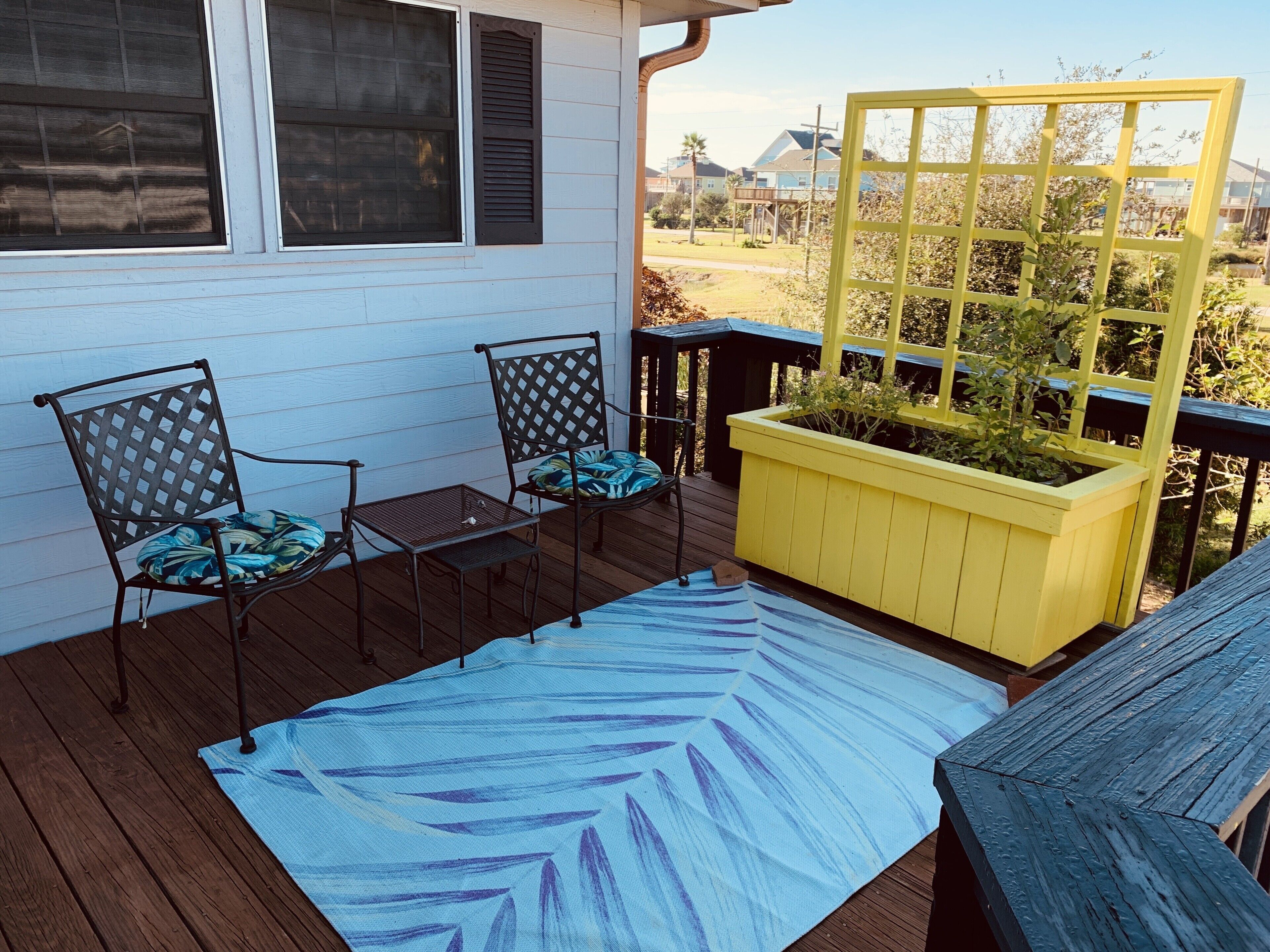 A deck with two chairs and a small table, a yellow planter box with plants, and a blue patterned rug.