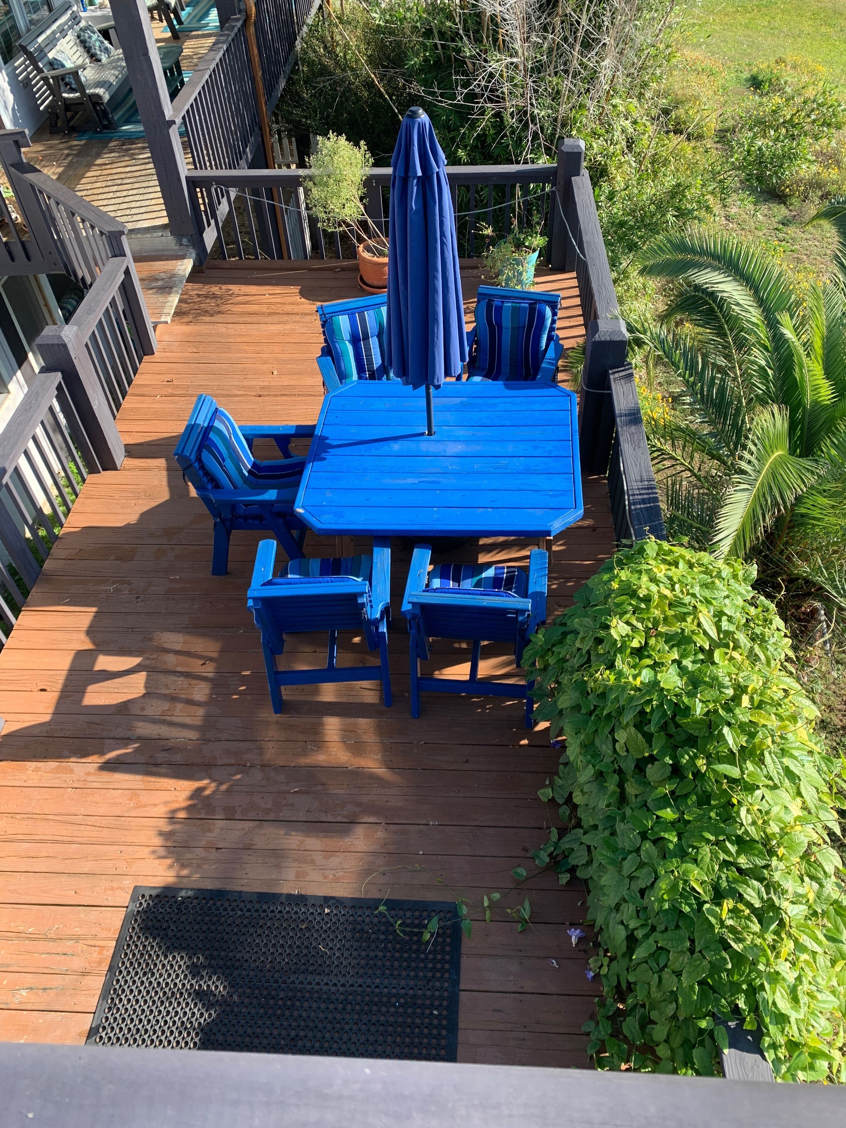 A wooden deck with a blue table and chairs, surrounded by greenery.