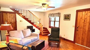 A living room with a staircase, wooden furniture, a ceiling fan, and a window with curtains. - Cottage on Mink River Road  (Ellison Bay)