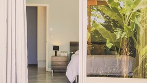A bedroom with a bed, bedside table, lamp, and a large window with a view of a banana tree. - Casa La Colina Oceanview Villa Santa Teresa Beach (Santa Teresa Beach)