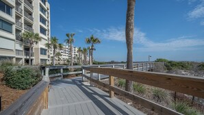 Una passerella di legno che conduce a una proprietà fronte spiaggia, con edifici di diverse altezze e palme.