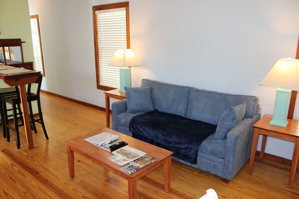 A living room with a sofa, a coffee table with magazines, a lamp, and a window with blinds.