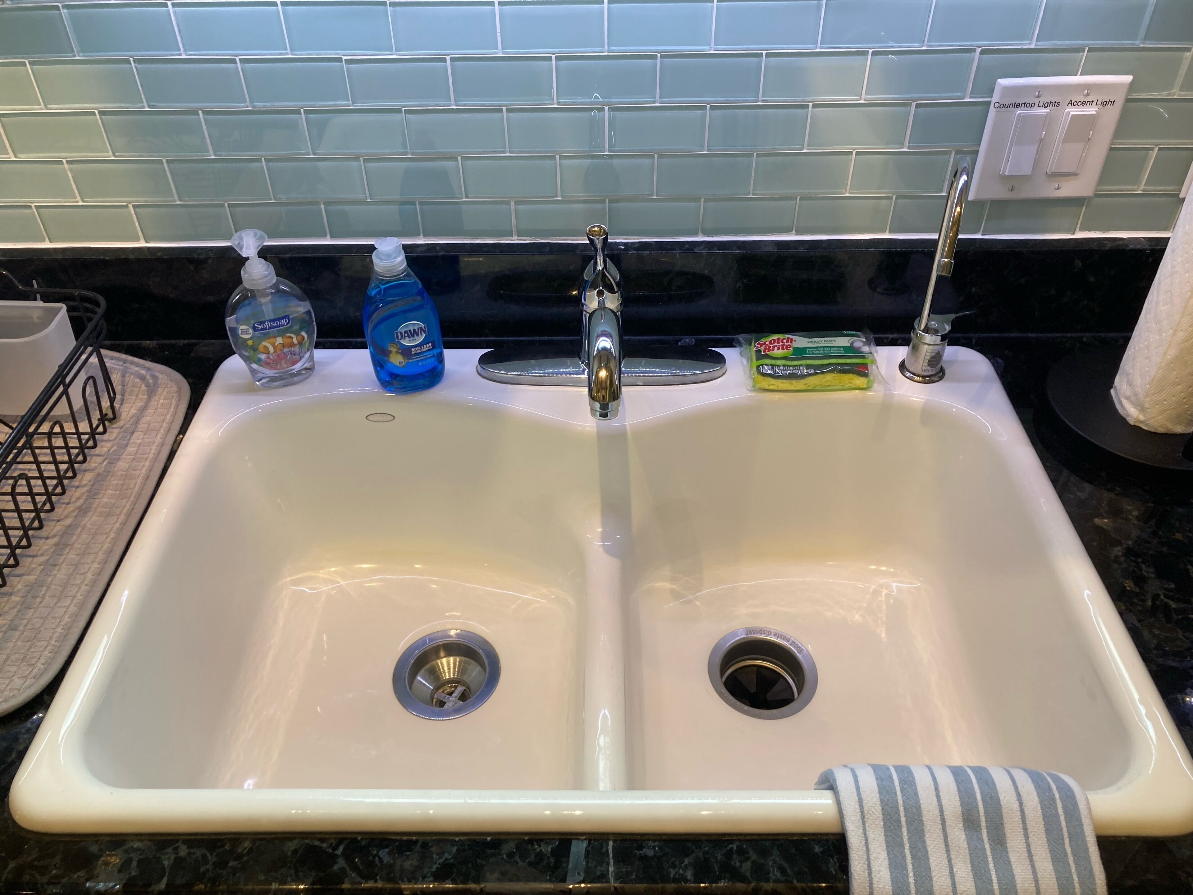 Ein Badezimmerwaschbecken mit zwei Wasserhähnen, einer Seifenspender, einer Flasche Dawn Geschirrspülmittel und einem Scotch-Brite Scheuerschwamm.