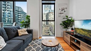 A modern living room with a grey sofa, a round coffee table, a TV on a wooden cabinet, and large windows with city views.
