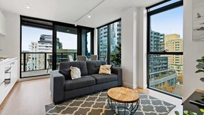A modern living room with a grey sofa, a round wooden coffee table, and large windows offering city views.