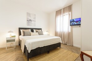 Quarto bem arrumado com cama, mesas de cabeceira, televisão e janela com cortinas.