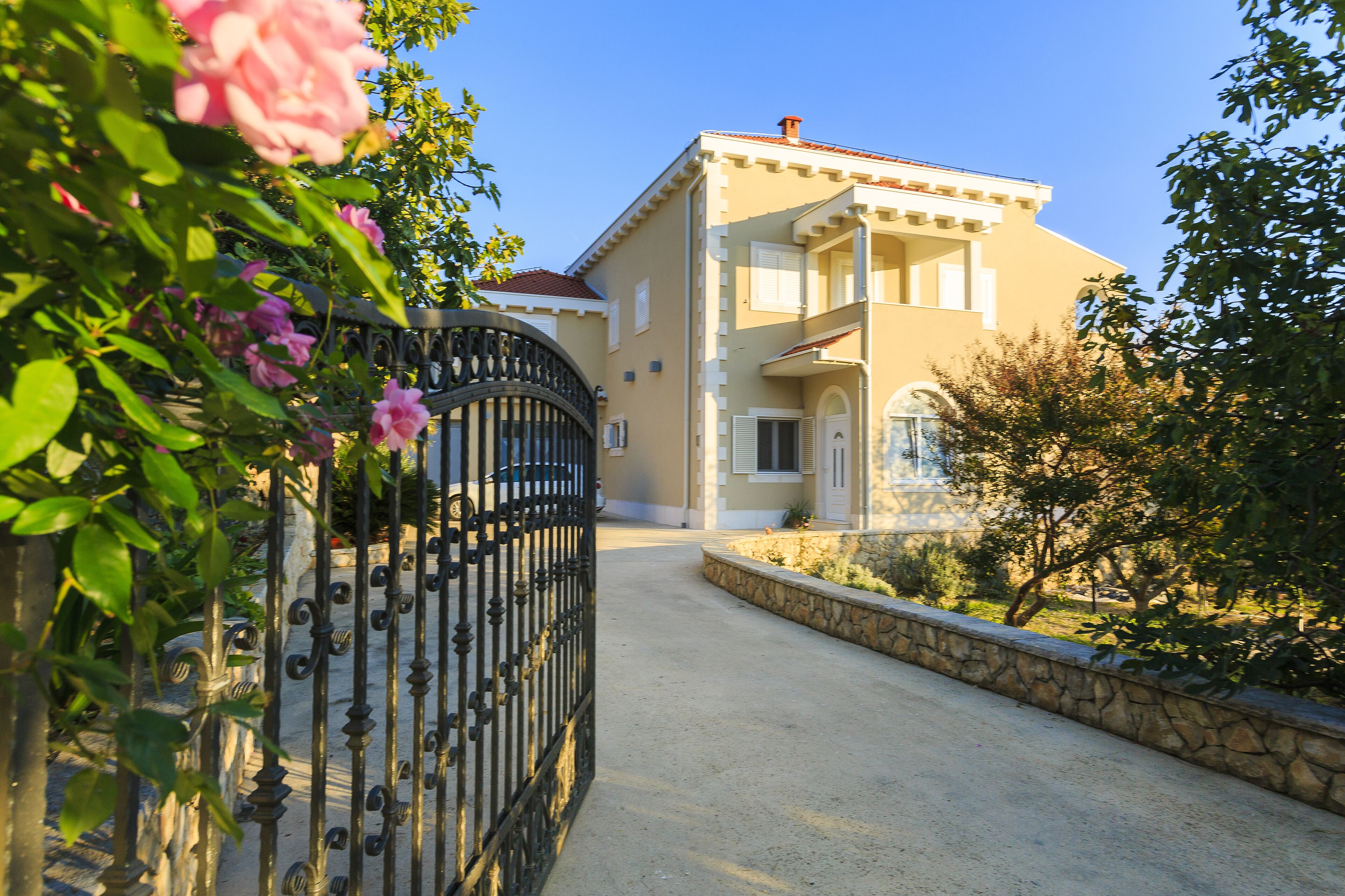 A two-story building with a stone pathway leading to the entrance, surrounded by greenery and a wrought iron gate.