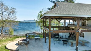 Une terrasse extérieure couverte, dotée d’un foyer en pierre, de tables et de chaises, et offrant une vue sur un lac.