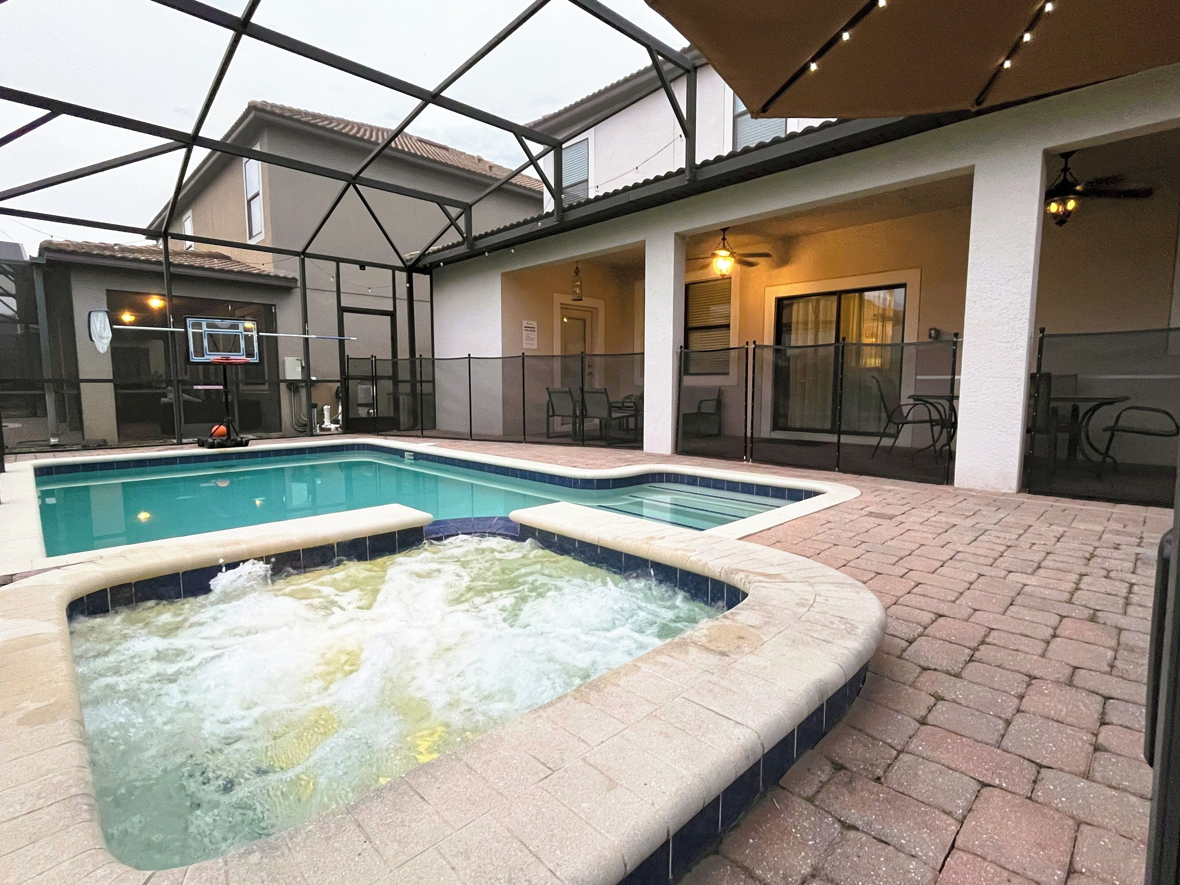 An indoor swimming pool with a hot tub, surrounded by a covered patio area with seating and a basketball hoop.