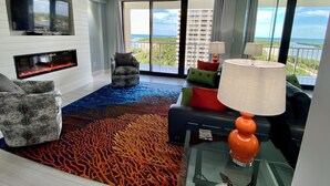 A modern living room with a flat-screen TV, a fireplace, a glass table, and a view of the ocean. - Newly Renovated Beach Condo with 2 Kings...Awesome Pool, Beach and Sunset Views (Marco Island)