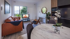 A modern living room with a sofa, TV, and dining area. - Jacaranda Apartment | Wayville Walk to Tram (Wayville)
