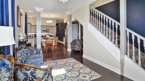 Un vaste salon agrémenté d’un canapé à motifs bleus, un coin repas avec une table et des chaises, une cuisine à l’arrière-plan et un escalier avec des balustrades blanches.