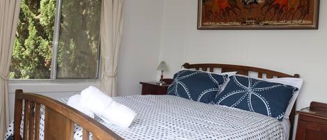 A bedroom with a wooden bed, a window with curtains, a nightstand, and a painting on the wall.