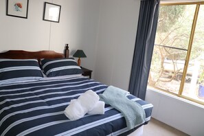 A neatly made bed with striped bedding, a bedside lamp, and a window with a view of trees and a vehicle outside. - Adrift at Ventnor, Pet Friendly on Phillip Island + wifi & linen (Ventnor)