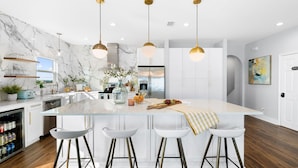 A modern kitchen with marble backsplash, white cabinetry, and a central island with bar stools. - Best Value Modern family friendly gulfront home (Gulf Shores)