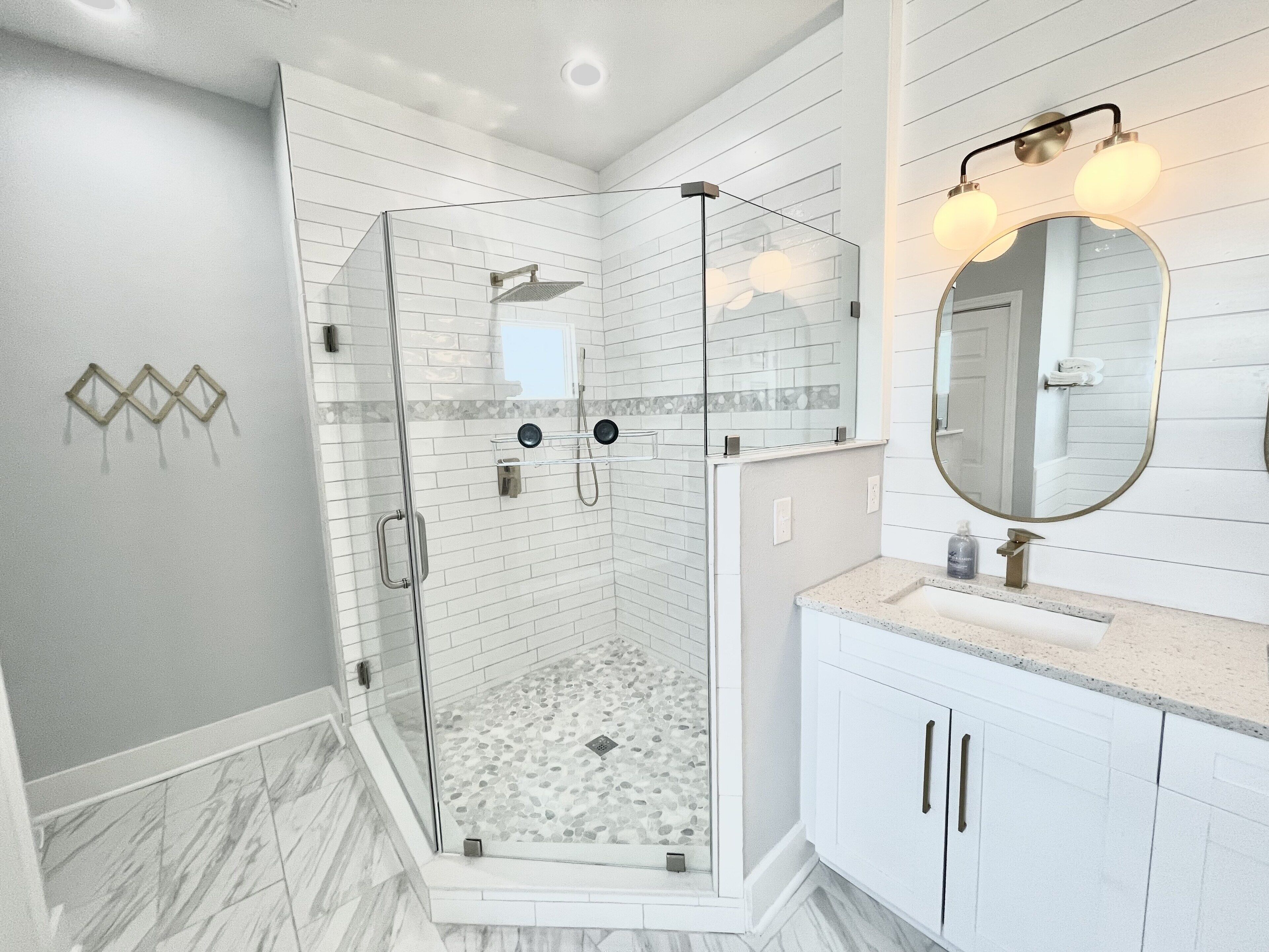 Un baño moderno con ducha de vidrio, paredes cubiertas de azulejos blancos, piso de mármol y un tocador con espejo e iluminación.