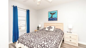 Quarto com cama, mesa de cabeceira, abajur, ventilador de teto e janela com cortinas.
