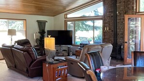 A spacious living room with a ceiling fan, a flat-screen TV, leather sofas, a dining table, and large windows offering a view of the outdoors. - Expansive Secluded Retreat w/ Hot tub, Mtn Views, 2mi to town, trails, birding (Leavenworth)