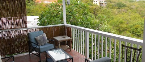 Ein Balkon mit Sitzgelegenheit, darunter eine Couch und Stühle, ein kleiner Tisch und Blick auf eine üppige grüne Landschaft mit Gebäuden im Hintergrund.