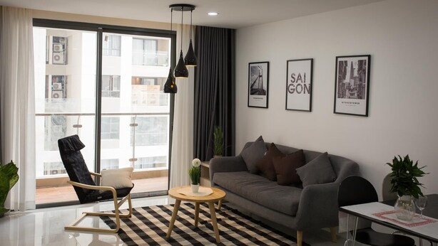 A modern living room with a sofa, a round coffee table, a black chair, and a dining table. There are three framed pictures on the wall, a large window with curtains, and a checkered rug. - Nice Apartment at GoldView (Ho Chi Minh City)