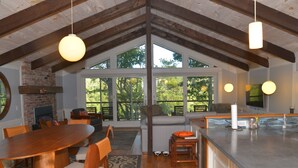 Amplia sala de estar con mesa de comedor de madera, sillas y sofá. Hay una chimenea, un ventanal grande y una zona de cocina con mostrador y lavamanos.
