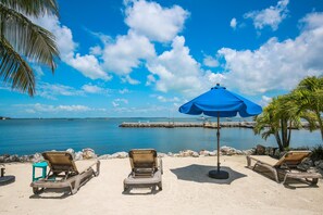 A beach with lounge chairs and a blue umbrella, a calm sea, and a clear sky.