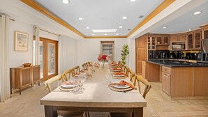 A spacious dining area with a long table set for a meal, wooden cabinets, and a kitchen area with modern appliances. - Elegant Home with Beach & Dockage! Spectacular Sunsets! (Key Largo)