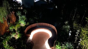 Un banco de madera curvo con una fuente de luz debajo, rodeado de plantas diversas y un camino de ladrillo.