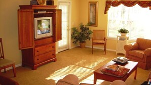 A living room with a television, wooden furniture, a ceiling fan, and a large window with curtains. - First Floor Luxury 1,350 Sq Ft  First Floor Condo w/ No Steps  (Myrtle Beach)