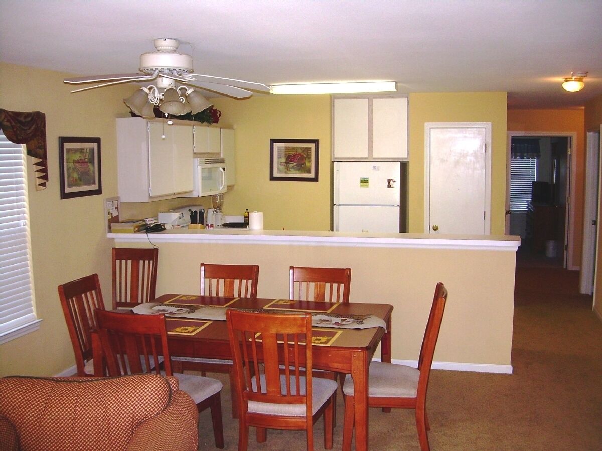 A dining area with a table, chairs, a ceiling fan, a kitchen area with cabinets and appliances, and a door leading to another room.