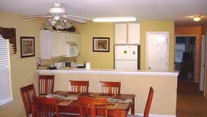 A dining area with a table, chairs, a ceiling fan, a kitchen area with cabinets and appliances, and a door leading to another room. - First Floor Luxury 1,350 Sq Ft  First Floor Condo w/ No Steps  (Myrtle Beach)