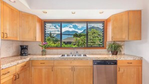 Una cocina con armarios de madera, lavavajillas de acero inoxidable, una ventana con vistas a montañas y vegetación, y una cafetera en la encimera.