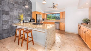 Cocina moderna con encimeras de granito, taburetes de madera y un amplio ventanal con vistas a un paisaje tropical.