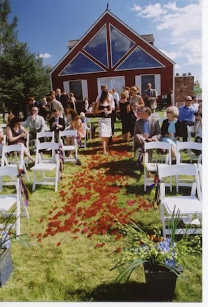Guests gather on lush green grounds for an intimate outdoor ceremony, surrounded by potted plants and a charming red building.