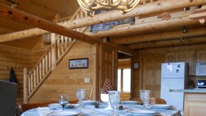 L’intérieur d’un chalet rustique en bois présente une table à manger pour quatre personnes, entourée de chaises en bois avec des coussins bleus. Un lustre est suspendu au plafond, et un escalier mène à un niveau supérieur.