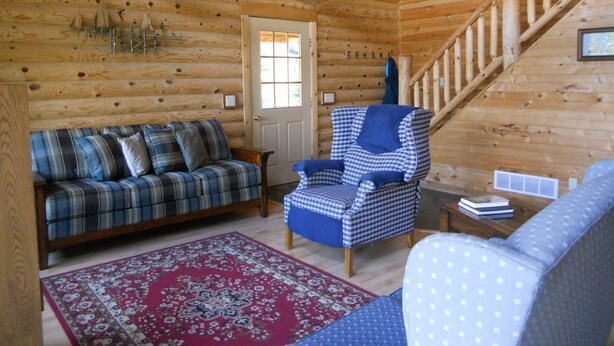 L’intérieur d’une cabane en rondins, avec un canapé à carreaux, un fauteuil à motifs, un tapis à dessins et un escalier en bois.