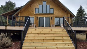 Une cabane en rondins avec un escalier en bois menant à une terrasse, une véranda couverte et une grande fenêtre.