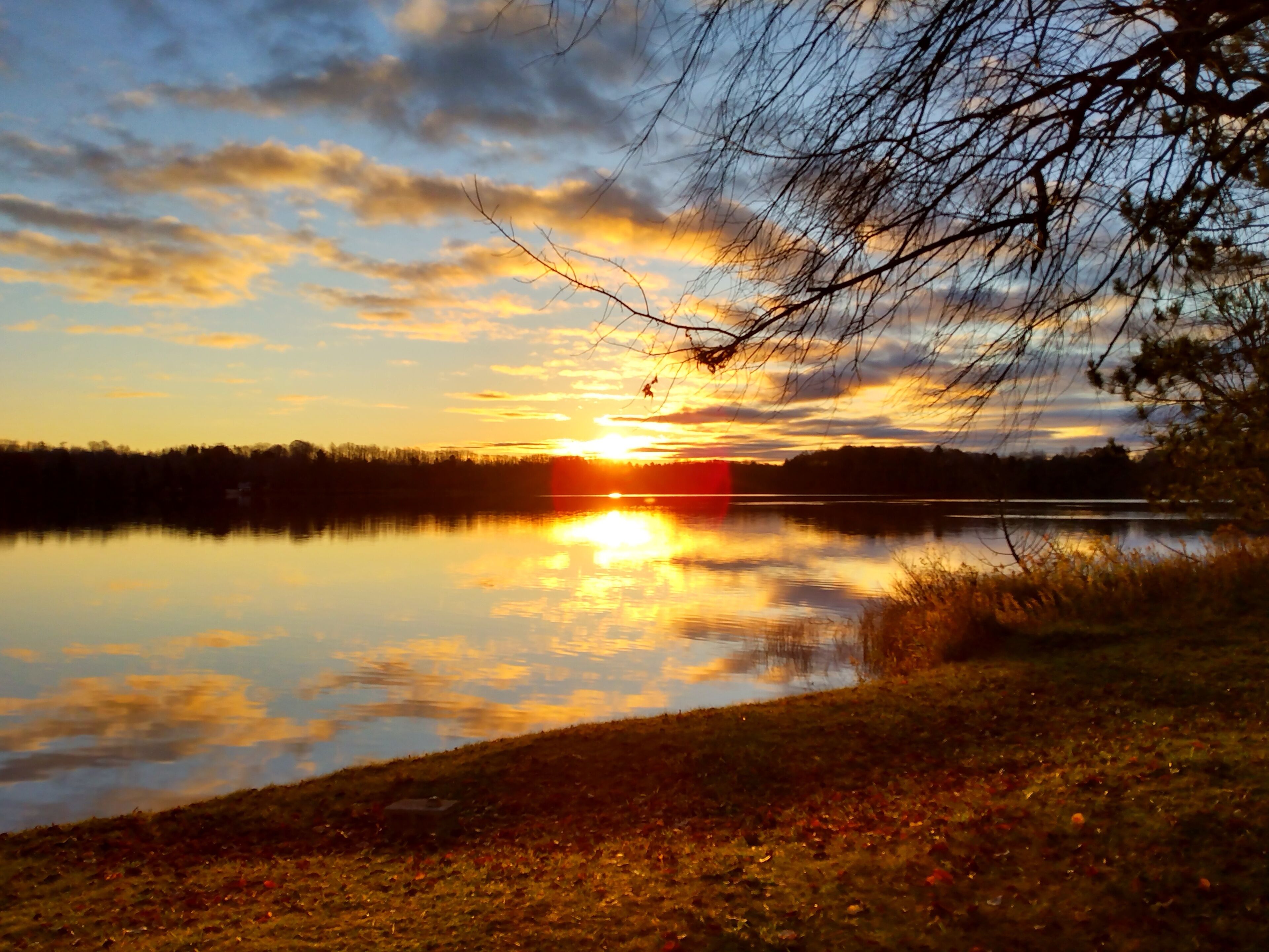 Un atardecer sobre un lago tranquilo con reflejos del cielo, rodeado de árboles y zonas verdes.