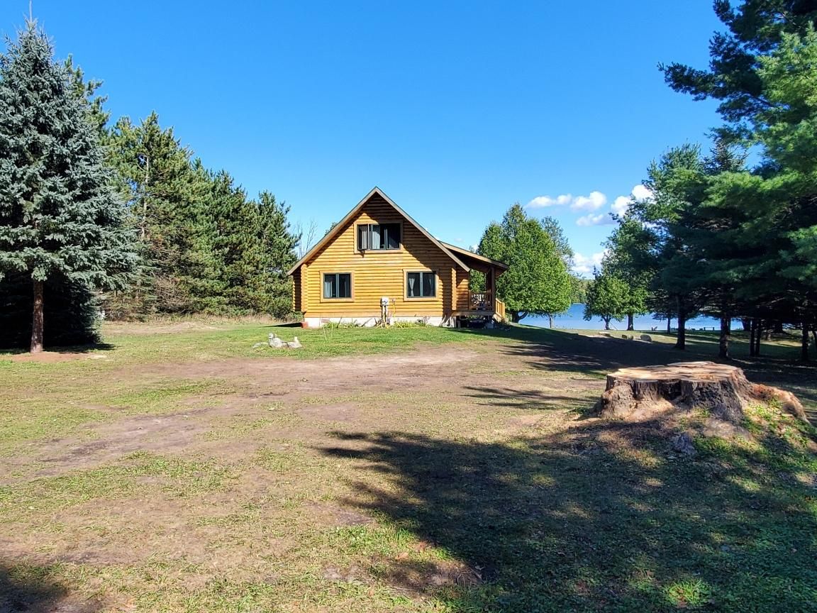 Une cabane en bois avec un toit à pignon, entourée d’une zone herbeuse et d’arbres, avec une étendue d’eau en arrière-plan.