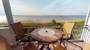 A balcony with a round table and wicker chairs, overlooking a beach with people and a boat in the distance.