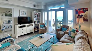 A spacious living room with a ceiling fan, a flat-screen TV, a bookshelf, wicker furniture, and a view of the ocean through large sliding glass doors.
