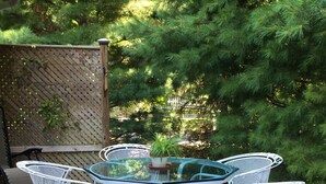 A round glass table with white metal chairs on a wooden deck, surrounded by green foliage. - WALK to Lake Michigan AND Saugatuck ~ Includes Bicycles! (Douglas)