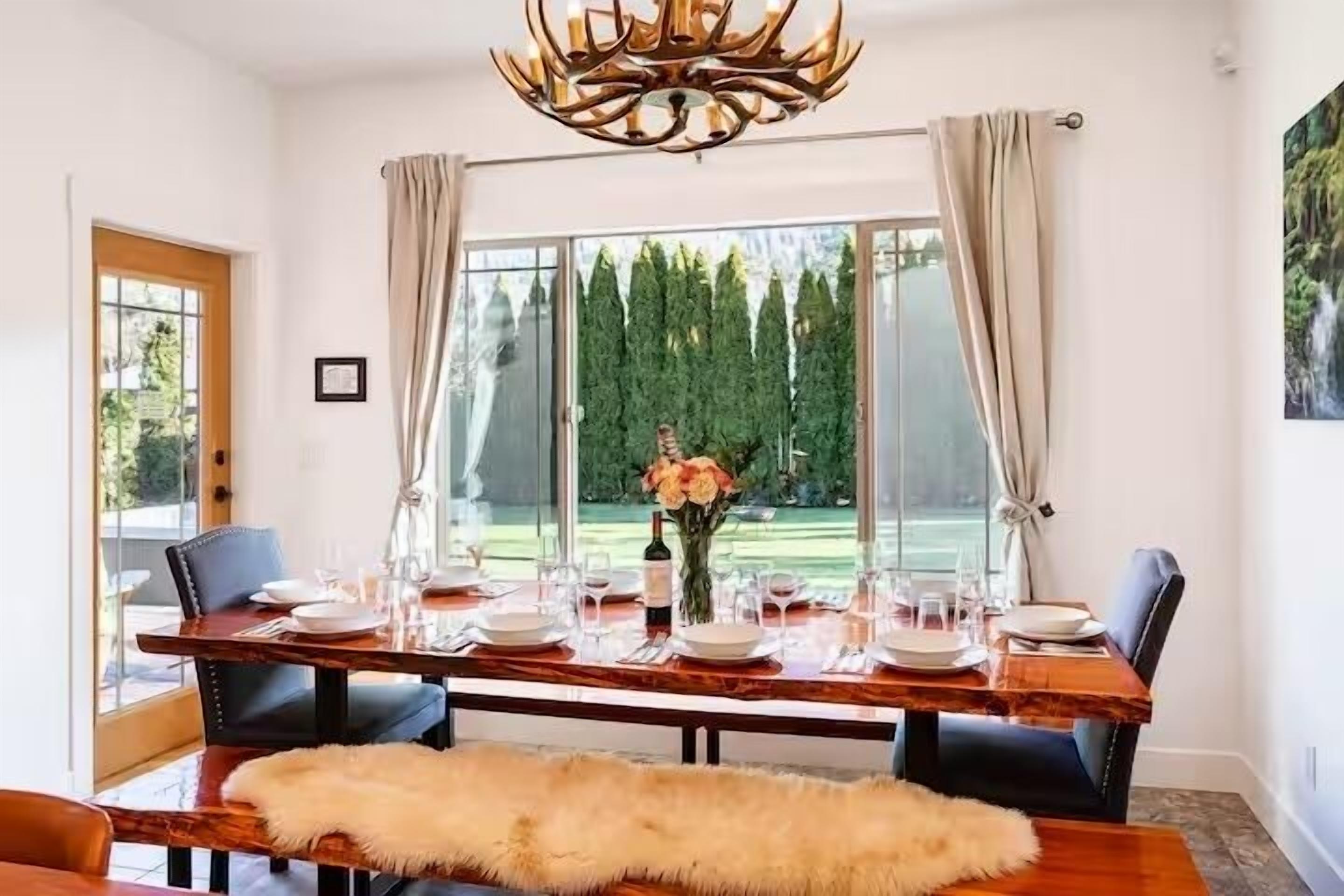 A dining area with a wooden table set for four, a chandelier, and a view of a garden through large windows.