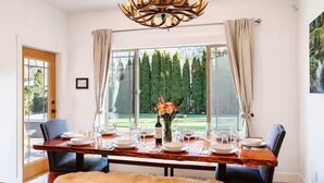 A dining area with a wooden table set for four, a chandelier, and a view of a garden through large windows.