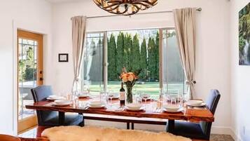 A dining area with a wooden table set for four, a chandelier, and a view of a garden through large windows.