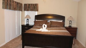 A neatly made bed with a swan-shaped towel, two bedside lamps, and a ceiling fan in a bedroom with a carpeted floor and curtains.