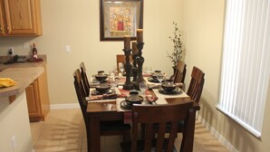 A dining area with a wooden table set for four, surrounded by chairs. A framed artwork hangs above the table, and a plant is visible to the right.